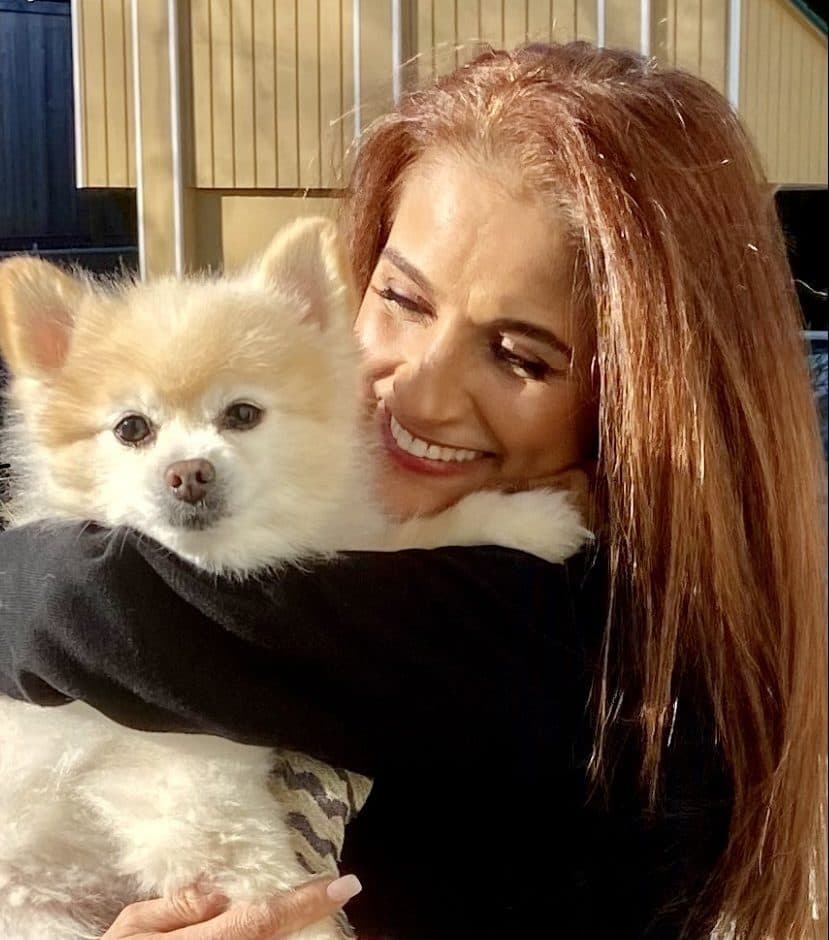 Soraya, with long auburn hair, smiling and hugging a small cream Pomeranian